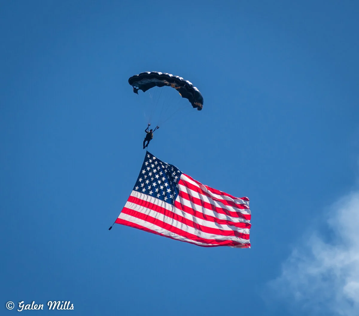 Parachutist descending with a large American flag against a blue sky.