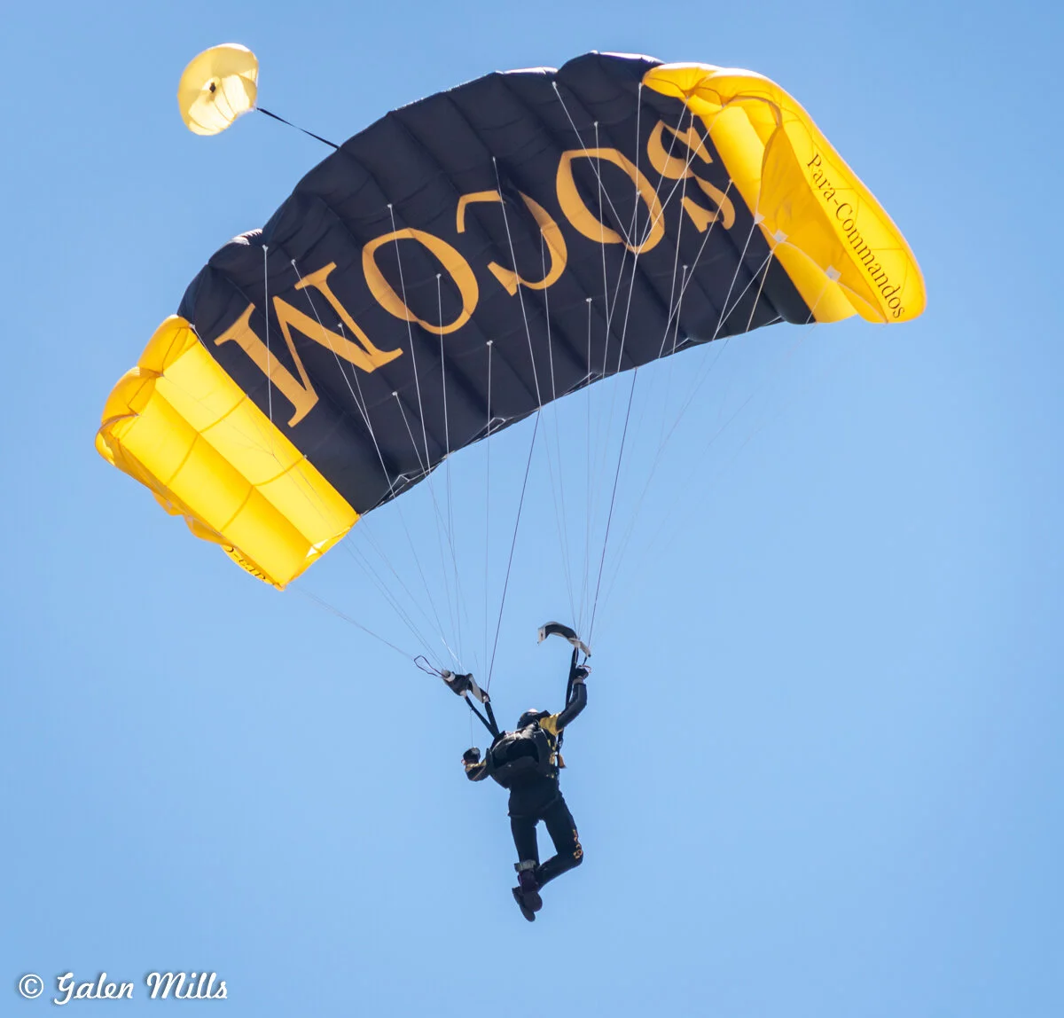 Skydiver descending with a black and yellow parachute, clear blue sky background.