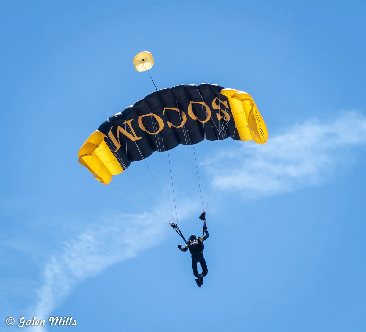 Skydiver with a black and yellow parachute against a clear blue sky.