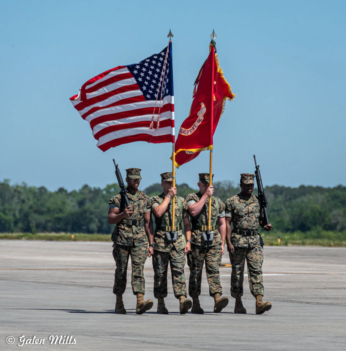 Four military personnel in uniform participate in a ceremonial march carrying the American flag and a red flag. Two additional personnel carry rifles. The background shows an open airfield and a line of trees.