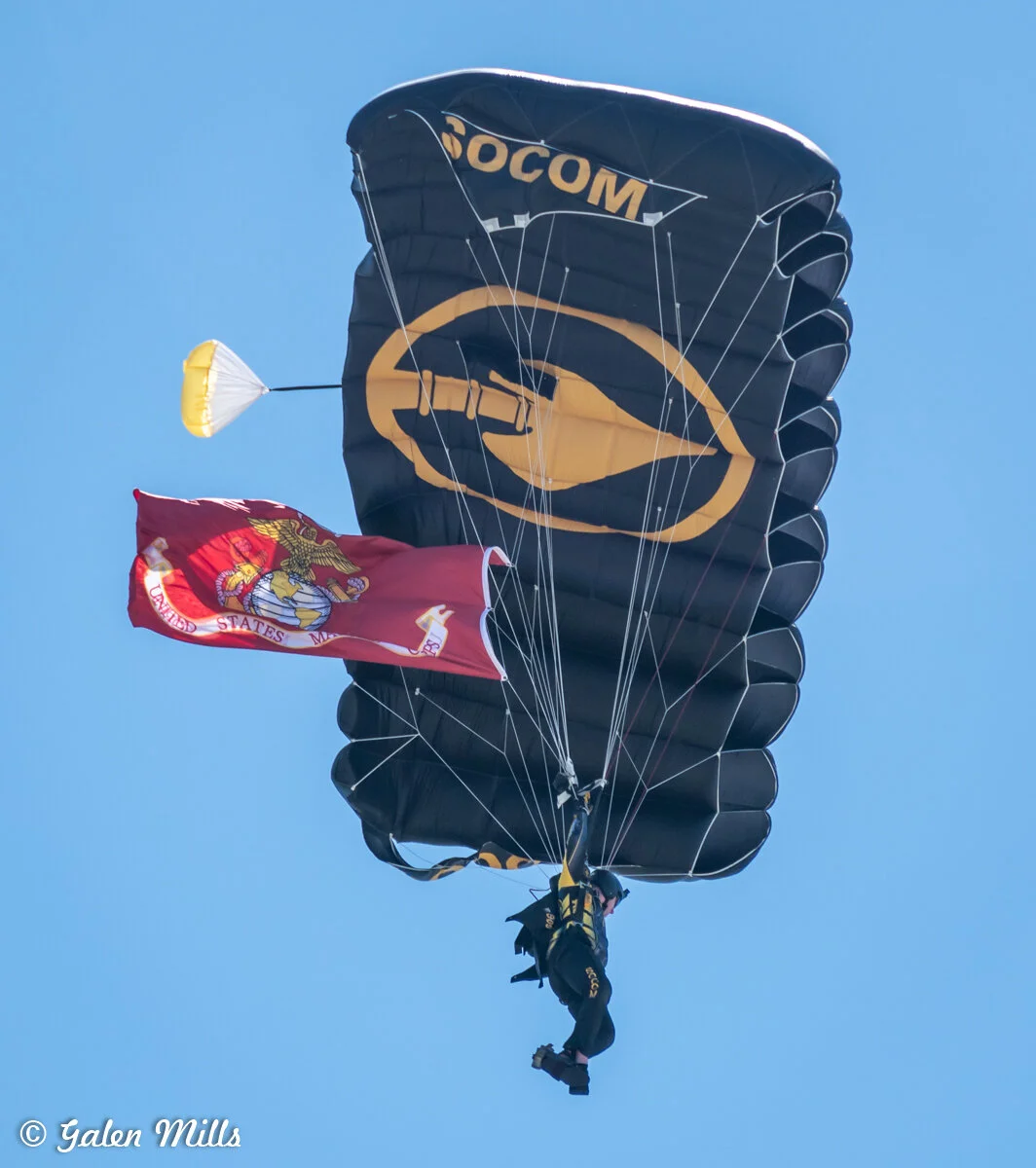 Parachutist descending with military-themed parachute and USMC flag.