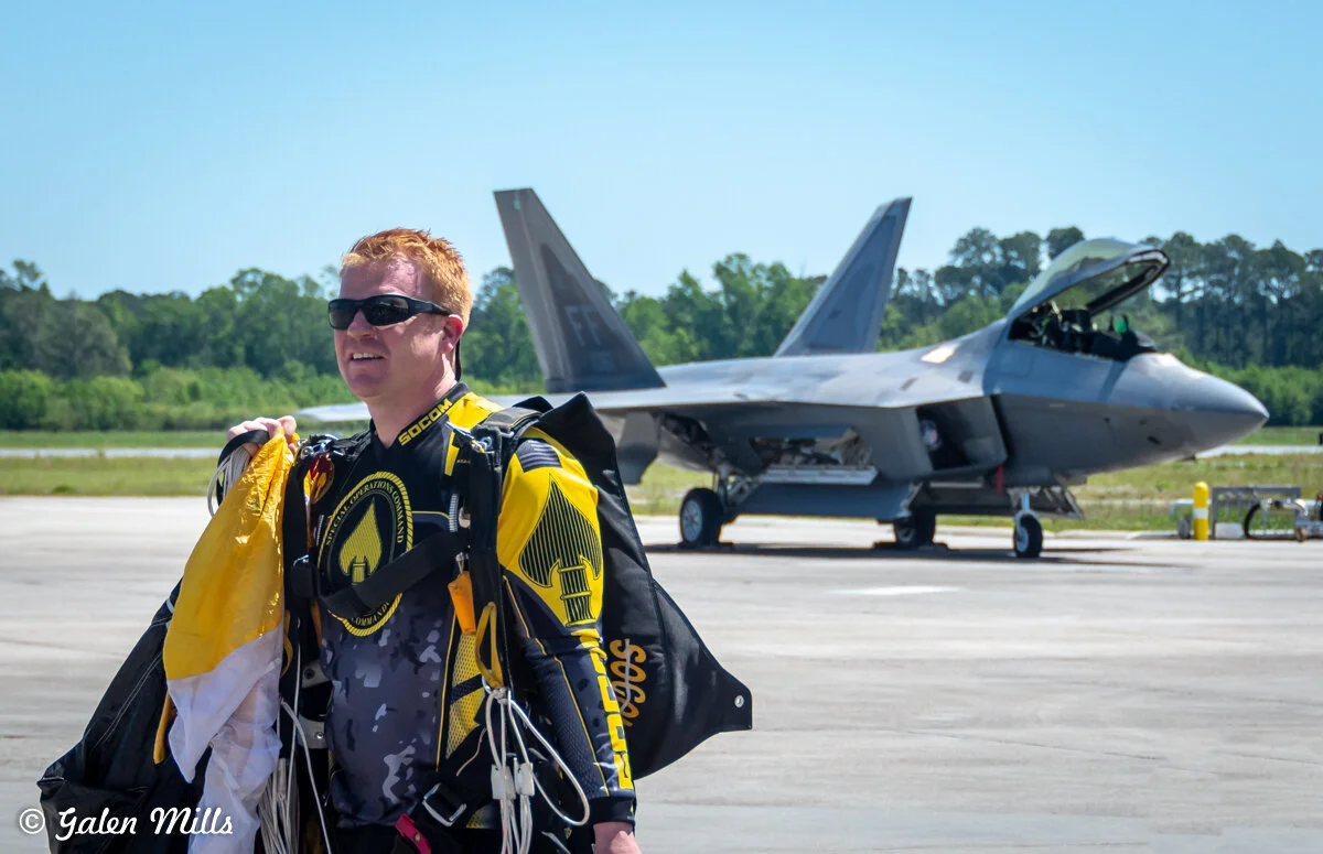 Skydiver in yellow and black gear on an airfield with a parked fighter jet in the background.