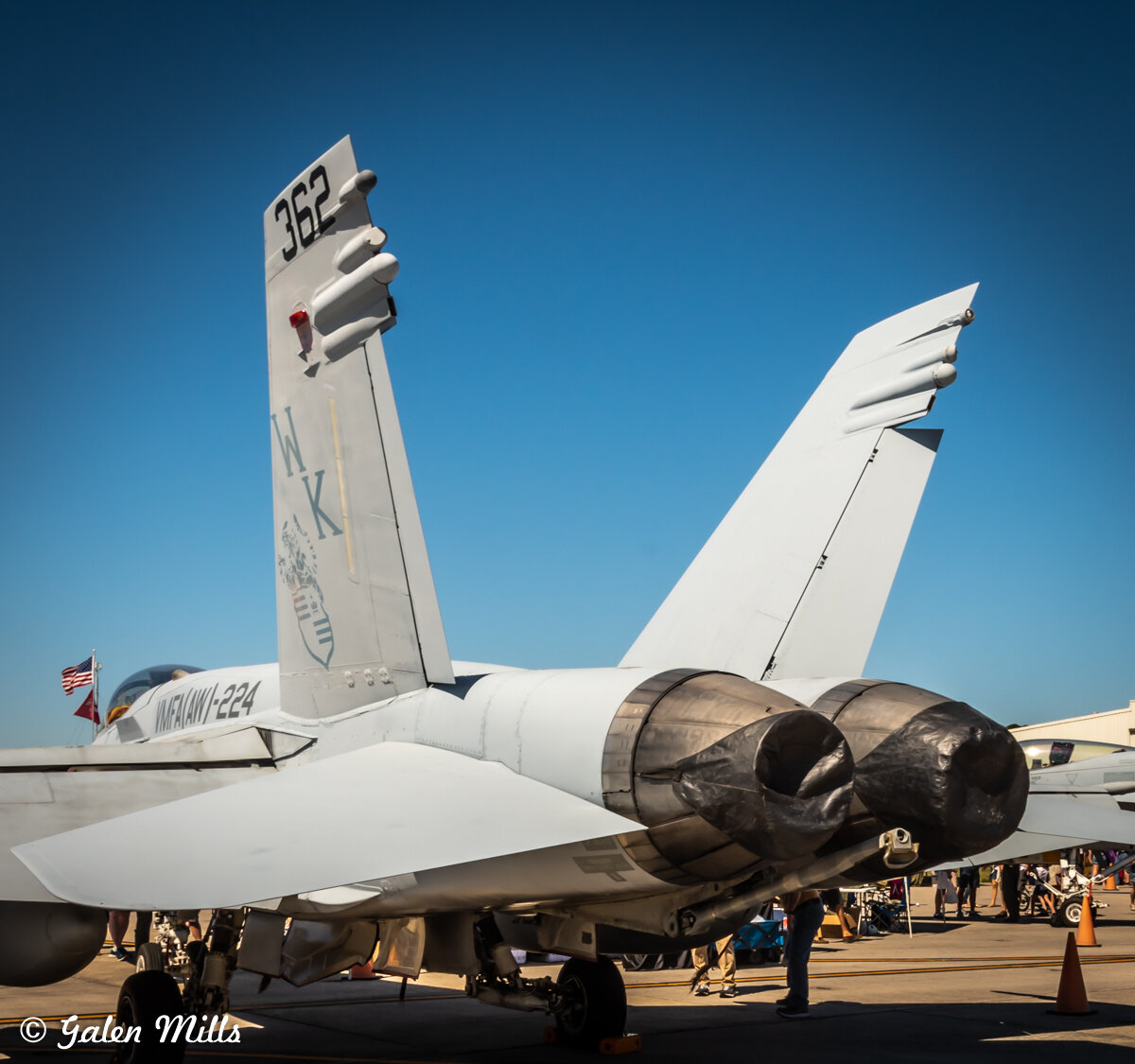Rear view of a military jet on display, showing twin exhaust nozzles and vertical stabilizers with markings, under a clear blue sky.