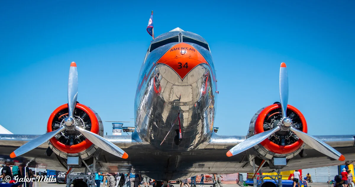 Front view of a shiny metallic vintage propeller airplane with two engines, set on an airport tarmac against a clear blue sky.