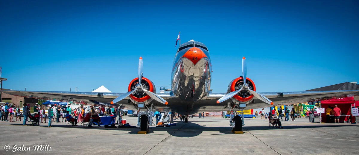 Front view of a vintage silver airplane with orange accents on display at an airshow, surrounded by people and tents.