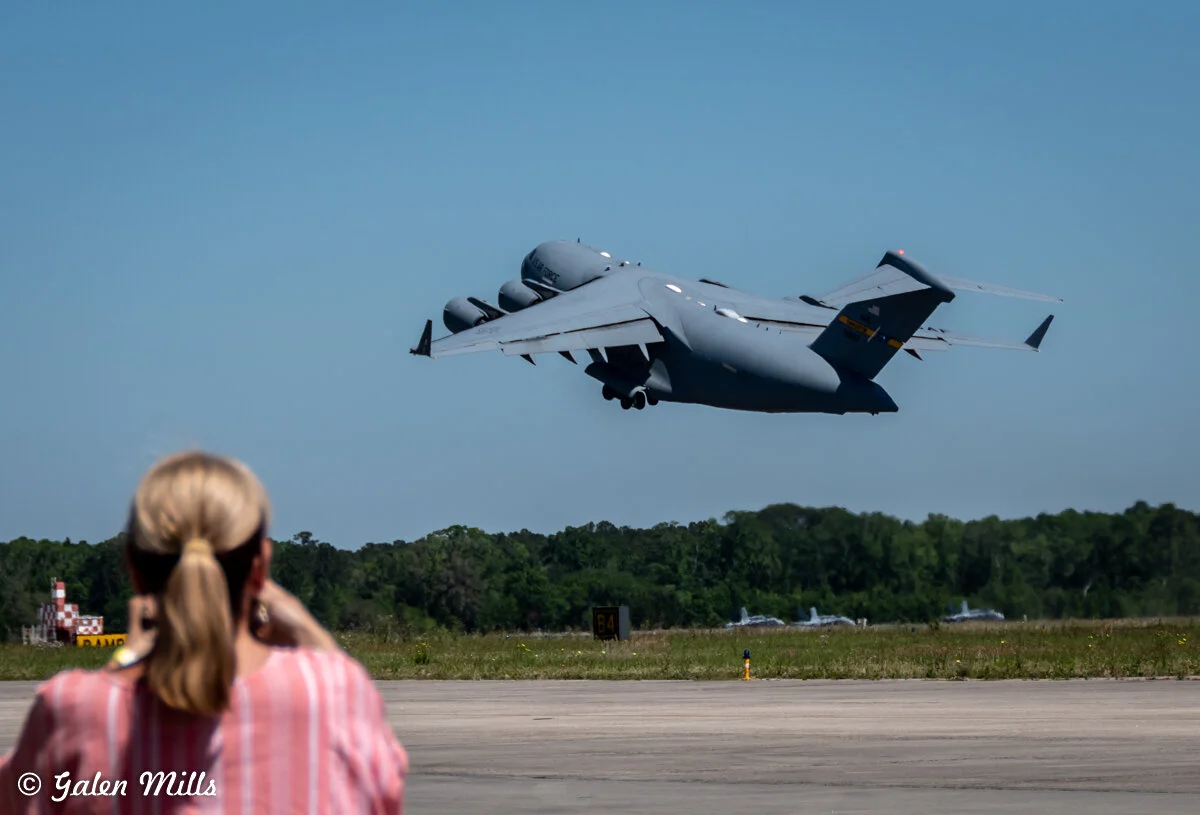 Person photographing a large military cargo plane taking off from an airstrip.
