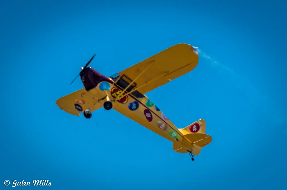 Yellow vintage airplane with colorful logos on its wings, flying against a clear blue sky.