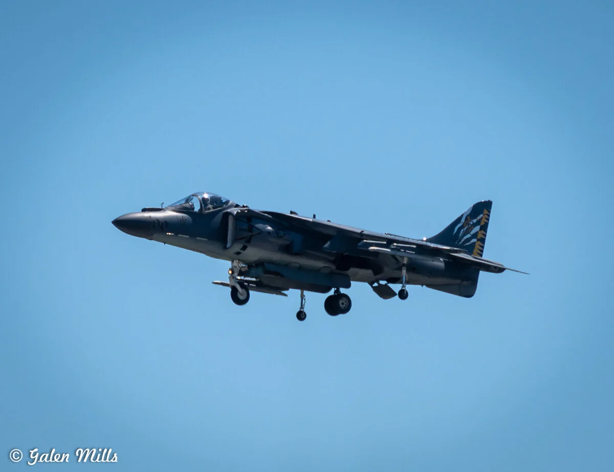A military jet aircraft flying with landing gear extended against a clear blue sky.