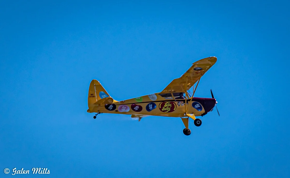 Yellow vintage airplane with Jelly Belly branding flying against a clear blue sky.
