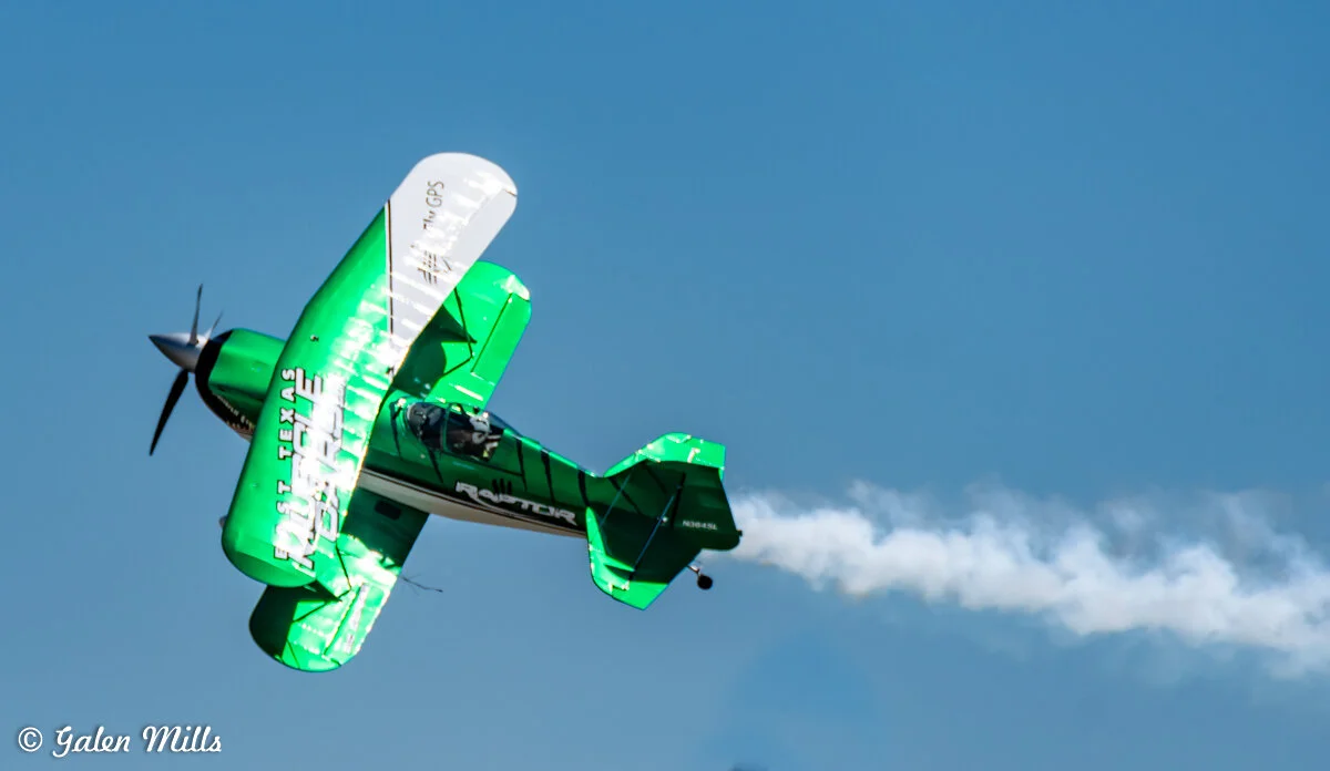 Green stunt biplane performing an aerobatic maneuver, emitting smoke trail against a clear blue sky.