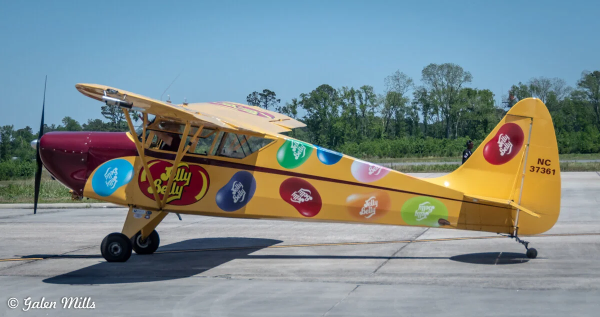 A small yellow and burgundy plane with Jelly Belly branding is parked on a runway. The plane features colorful jelly bean graphics and decals with the Jelly Belly logo.