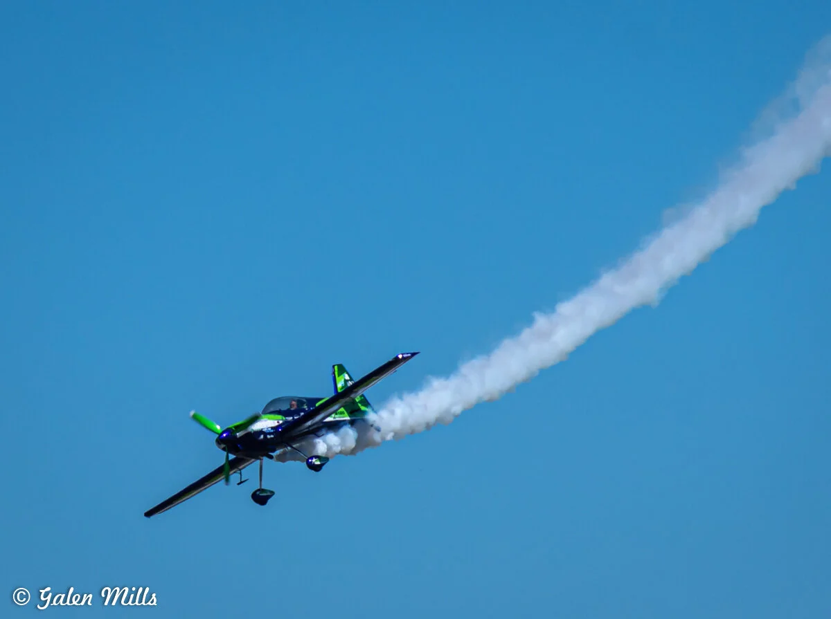 Aerobatic plane in flight with smoke trail against clear blue sky.