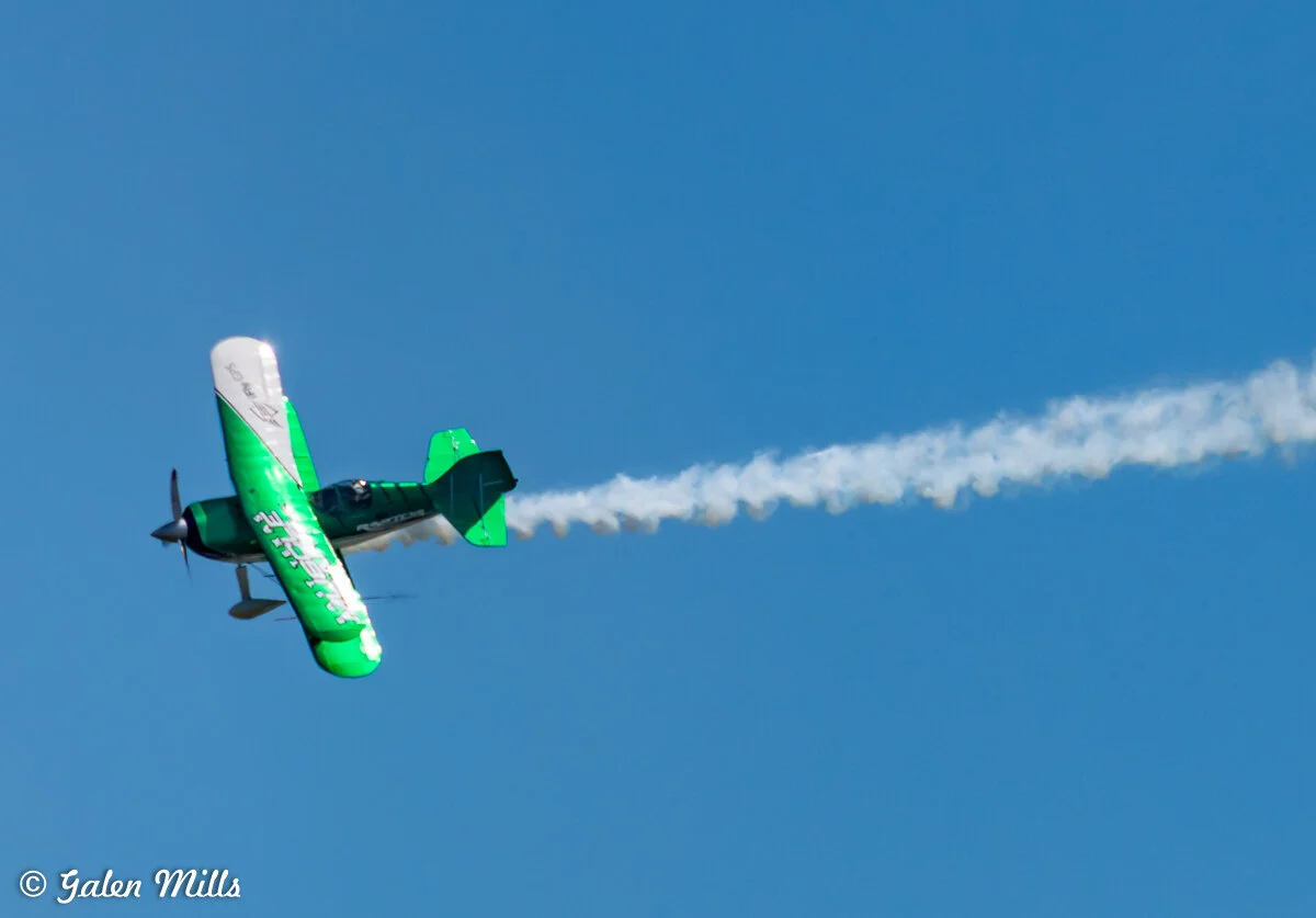 Green and white biplane performing aerobatic maneuver with smoke trail against clear blue sky.