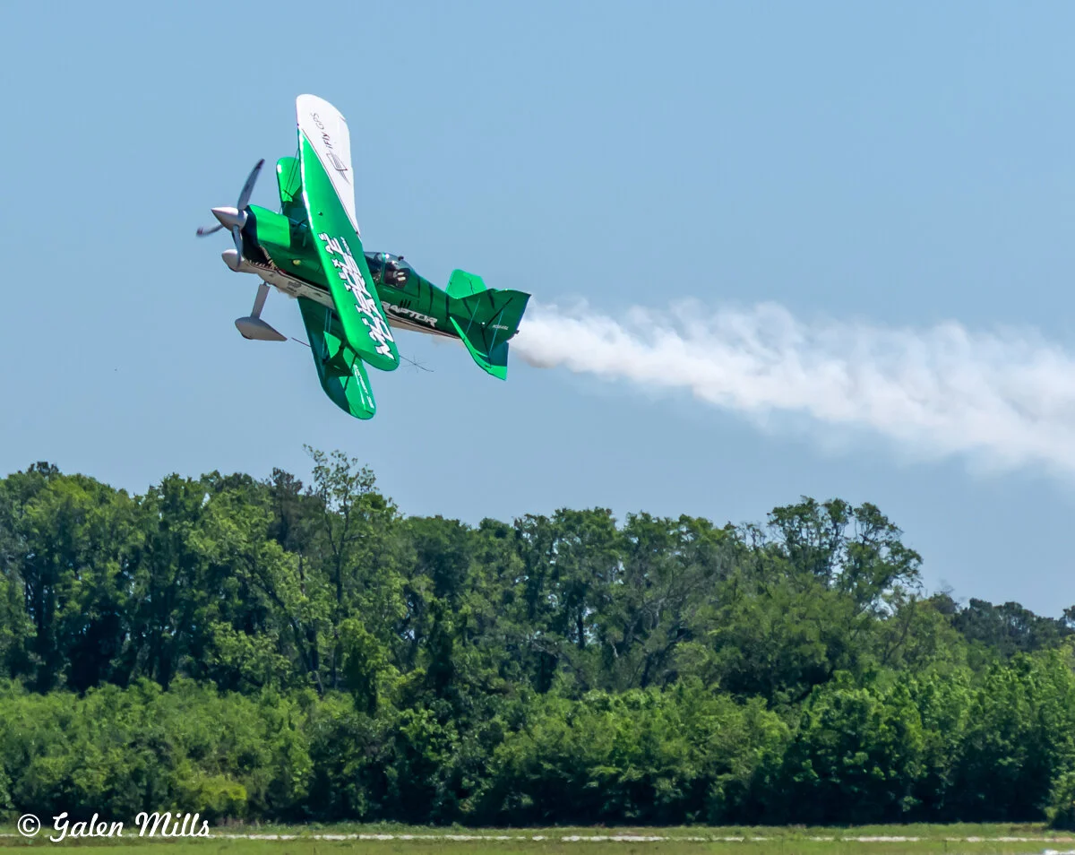 Green stunt plane performing an aerobatic maneuver with smoke trailing, set against a clear blue sky and green treeline backdrop.