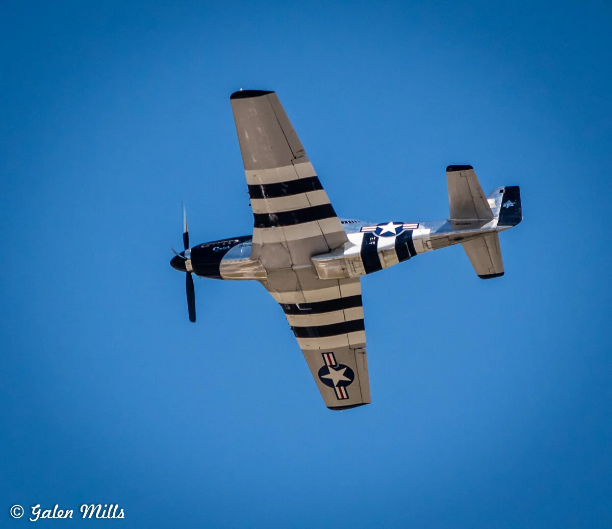 A vintage military aircraft with black and white stripes flying against a clear blue sky.