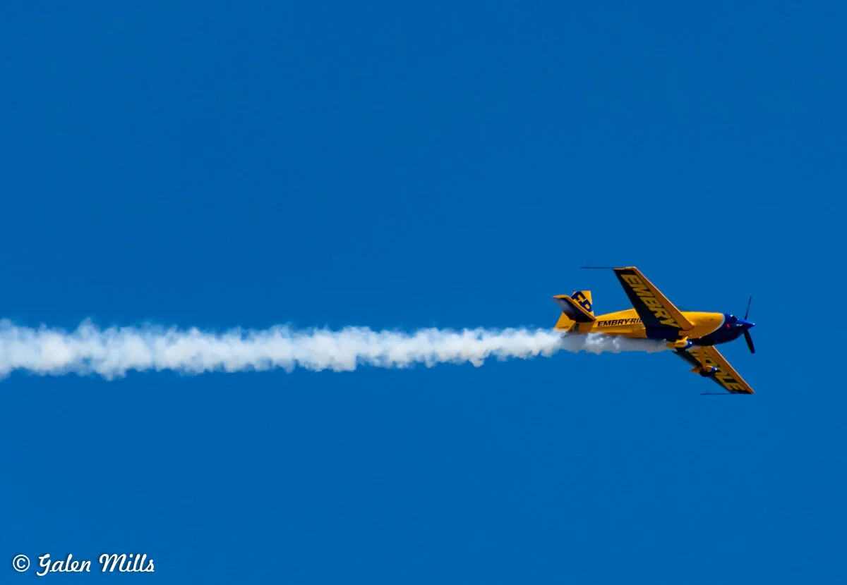 Yellow aerobatic airplane performing skywriting with smoke trail against a clear blue sky.