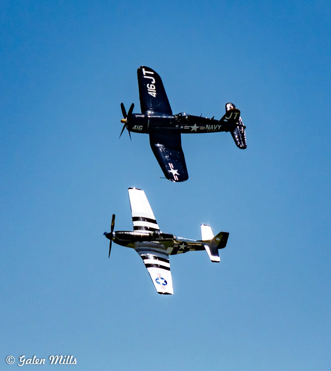 Two vintage military airplanes fly in formation in a clear blue sky, featuring distinct navy and white markings.