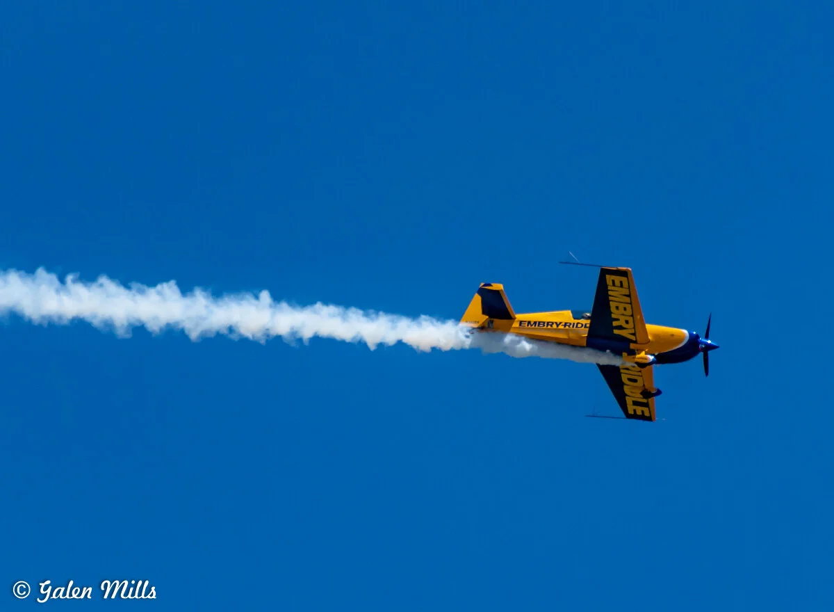 Yellow and blue aerobatic airplane with "Embry-Riddle" on the wings, performing a stunt with smoke trail against a clear blue sky.