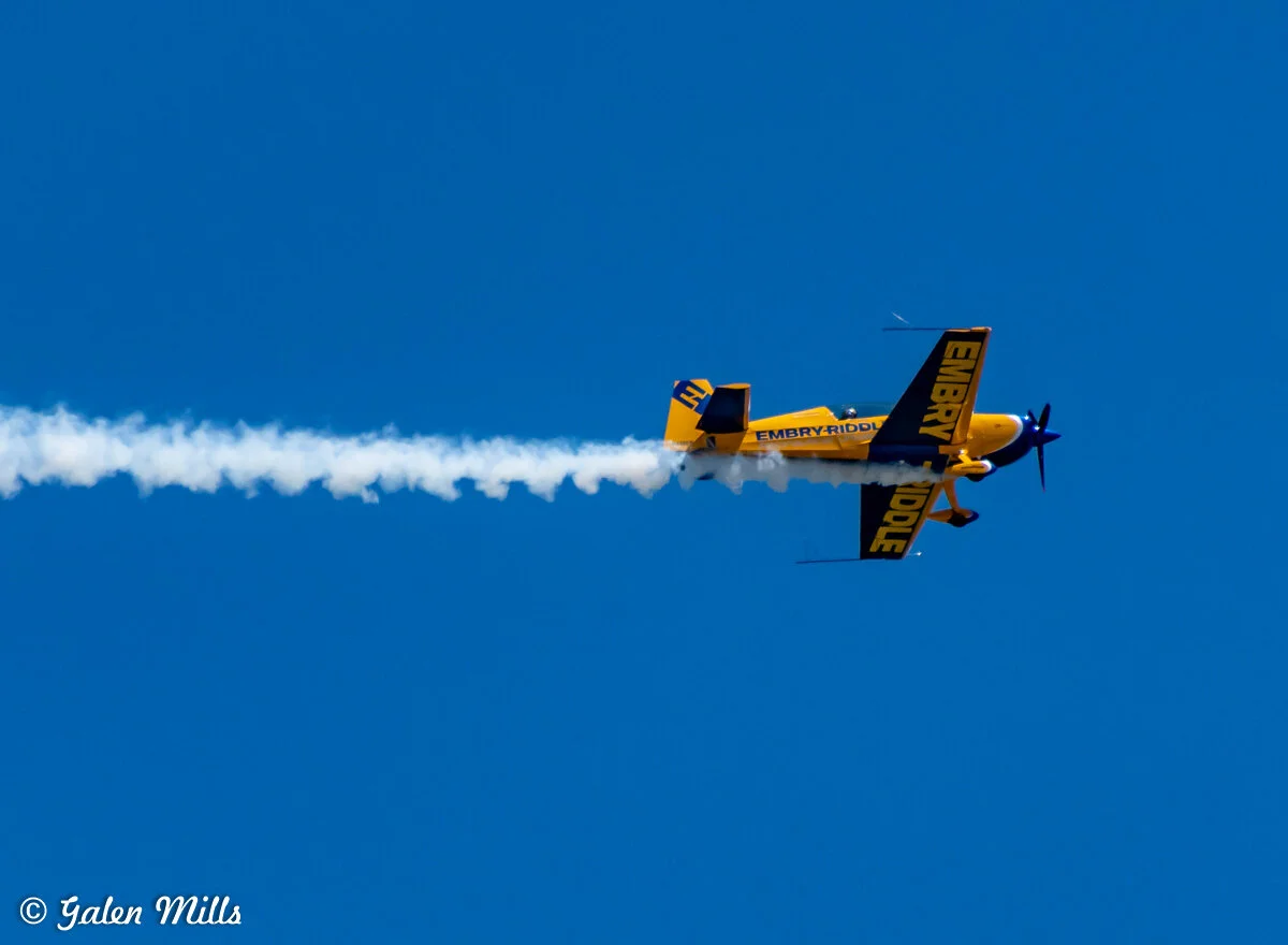 Aerobatic airplane from Embry-Riddle performing a maneuver with smoke trail against a clear blue sky.