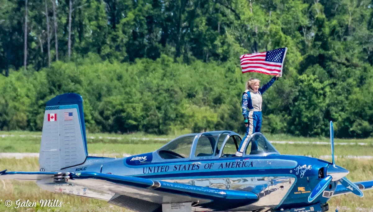 A person in a jumpsuit stands on a blue aircraft, holding a waving American flag, with trees in the background.
