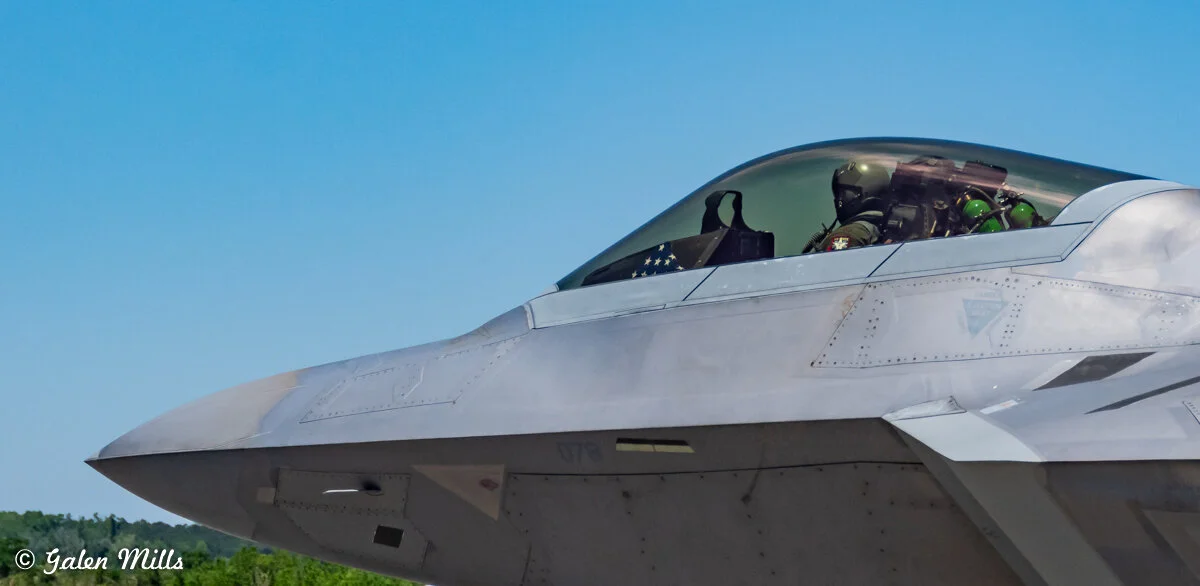 Close-up of a modern fighter jet cockpit with a pilot inside, set against a clear blue sky.