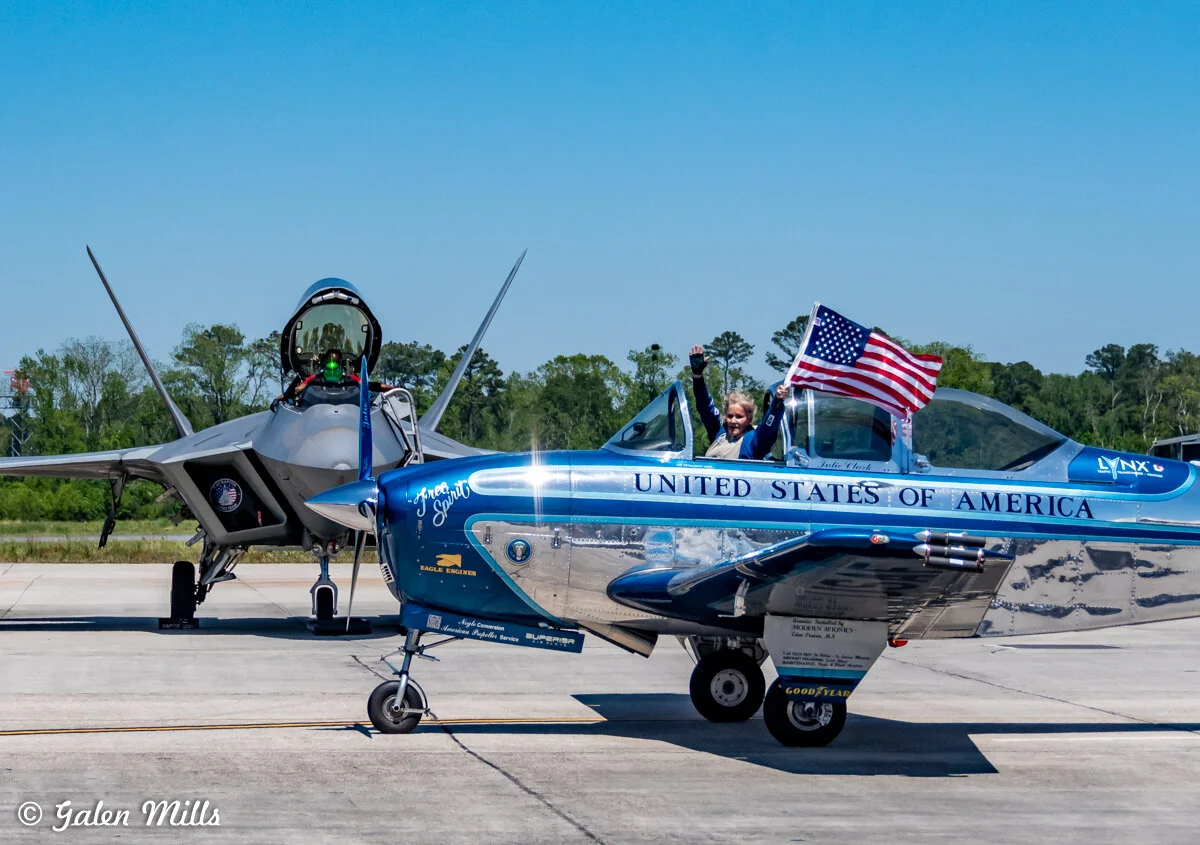 A small aircraft with "United States of America" painted on the side and an American flag being waved by a woman. An F-22 Raptor jet is parked in the background on an airfield.