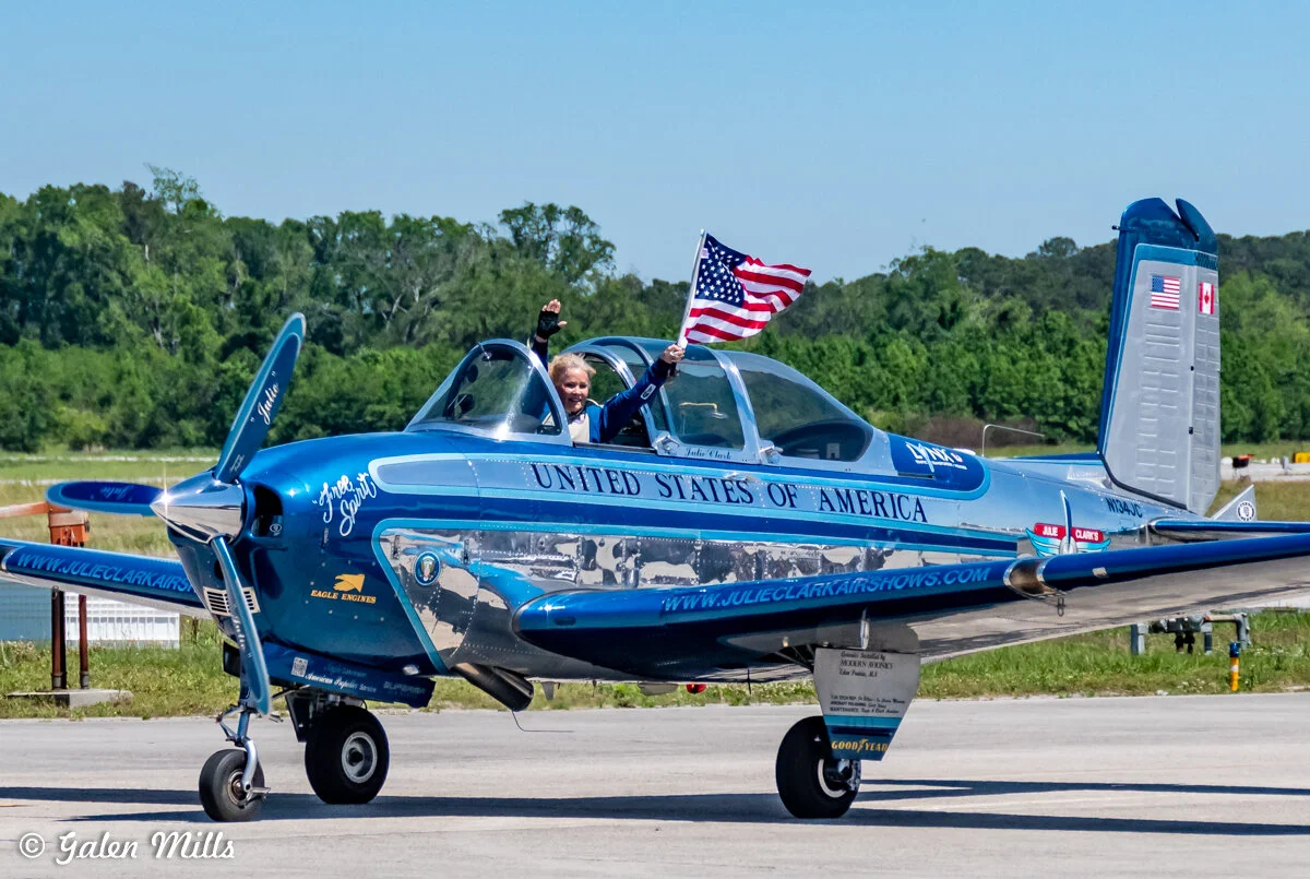 A blue aircraft with "United States of America" written on the side is on a runway. A person is waving an American flag from the cockpit. The tail features both American and Canadian flags. The plane's propeller is stationary.