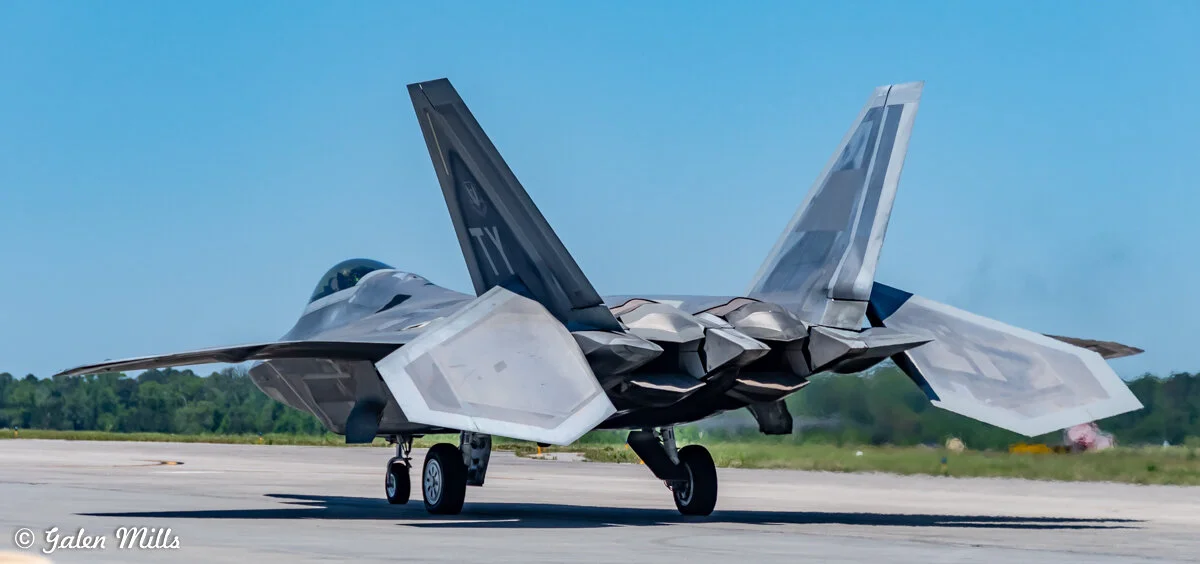Fighter jet taxiing on a runway under clear blue sky. The view is from the rear, showing the jet's wings and tail.