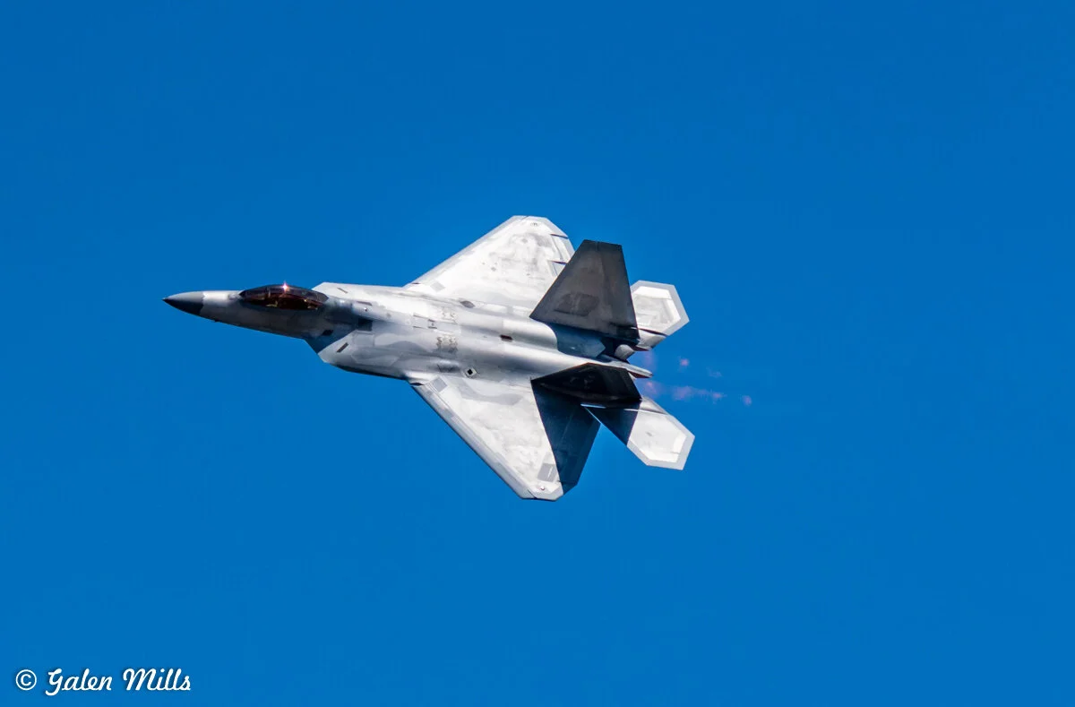 A military jet aircraft flying in a clear blue sky with visible vapor trails.
