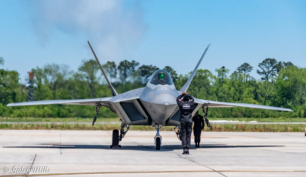 Military jet with open canopy on tarmac and personnel nearby with forest background.