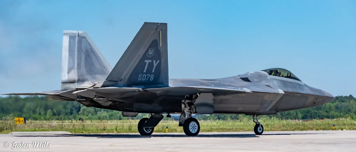 F-22 Raptor fighter jet on a runway with landing gear extended, bearing markings "TY" and "AF 04 078" on the tail, set against a clear sky and countryside background.