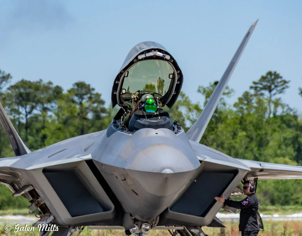 A military fighter jet with open cockpit and a pilot inside, positioned on a runway with a crew member nearby, wearing headphones. Trees are visible in the background.