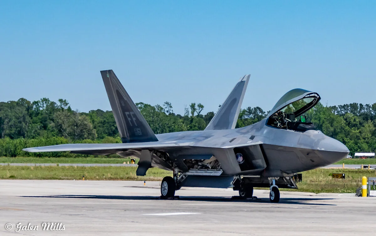 F-22 Raptor fighter jet parked on a tarmac with open cockpit, trees in the background.