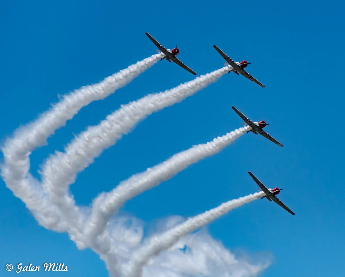 Four planes flying in formation, leaving smoke trails, against a blue sky.