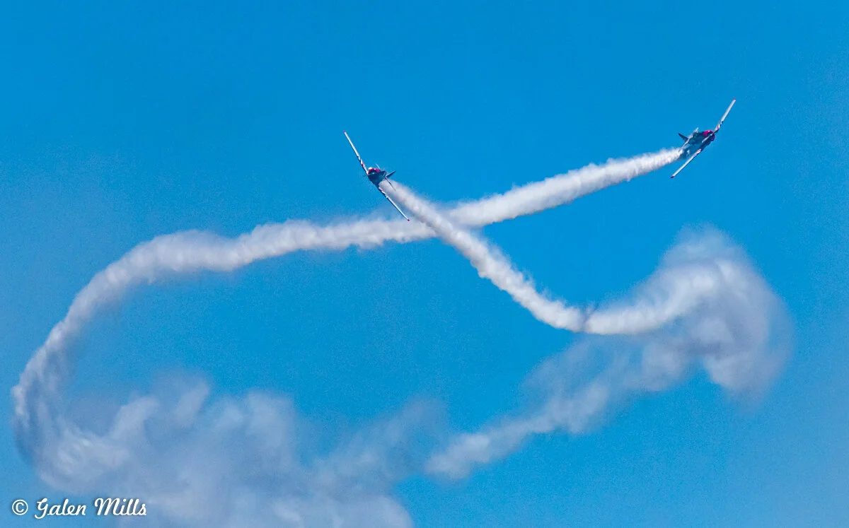 Two airplanes performing aerial maneuvers with smoke trails against a blue sky.