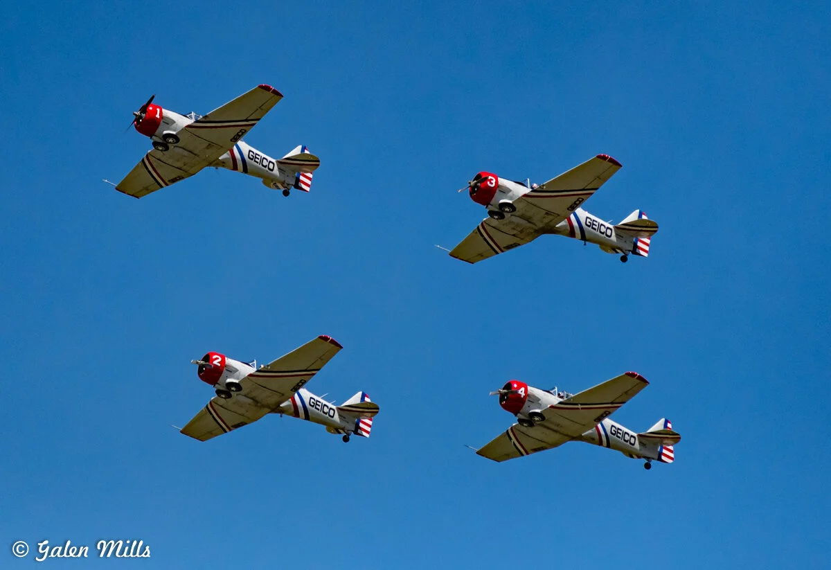 Four vintage military aircraft flying in formation against a clear blue sky, with "GEICO" branding visible on the fuselage.