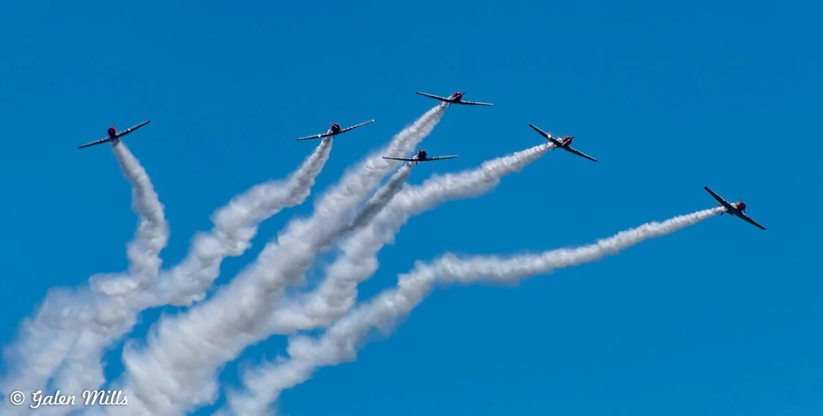 Formation of five airplanes emitting smoke trails against a clear blue sky.