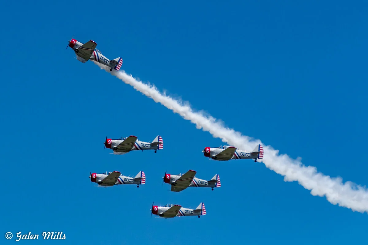 A formation of six vintage propeller aircraft performing an aerial display against a clear blue sky, trailing smoke in synchronized maneuvers. The aircraft bear the logo and colors of GEICO.