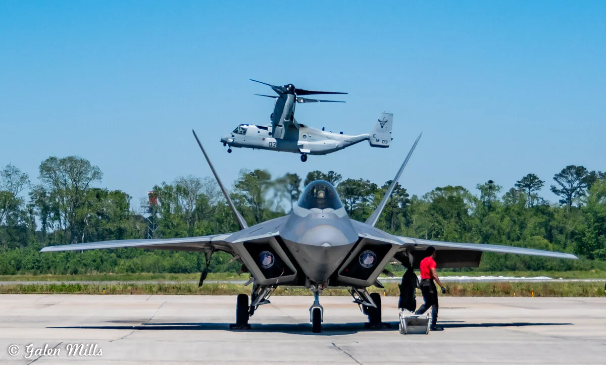 A military jet and a tiltrotor aircraft on a runway with trees in the background, under a clear sky. A person in a red shirt is standing near the jet.