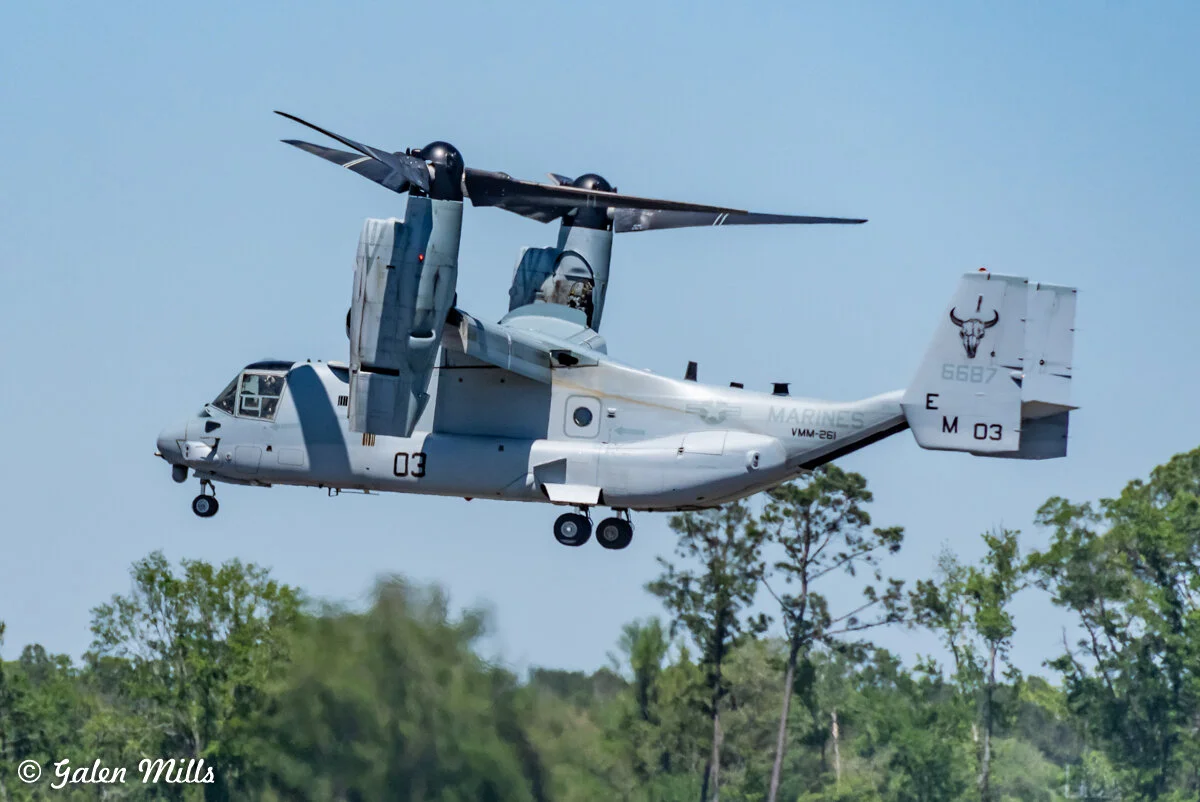 V-22 Osprey tiltrotor aircraft in flight with trees in the background.