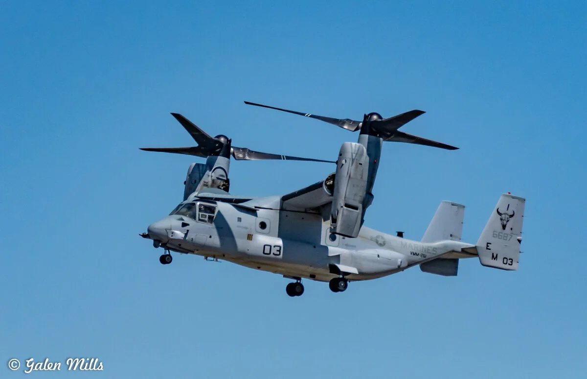 A military V-22 Osprey aircraft in flight with rotors in horizontal position against a clear blue sky.