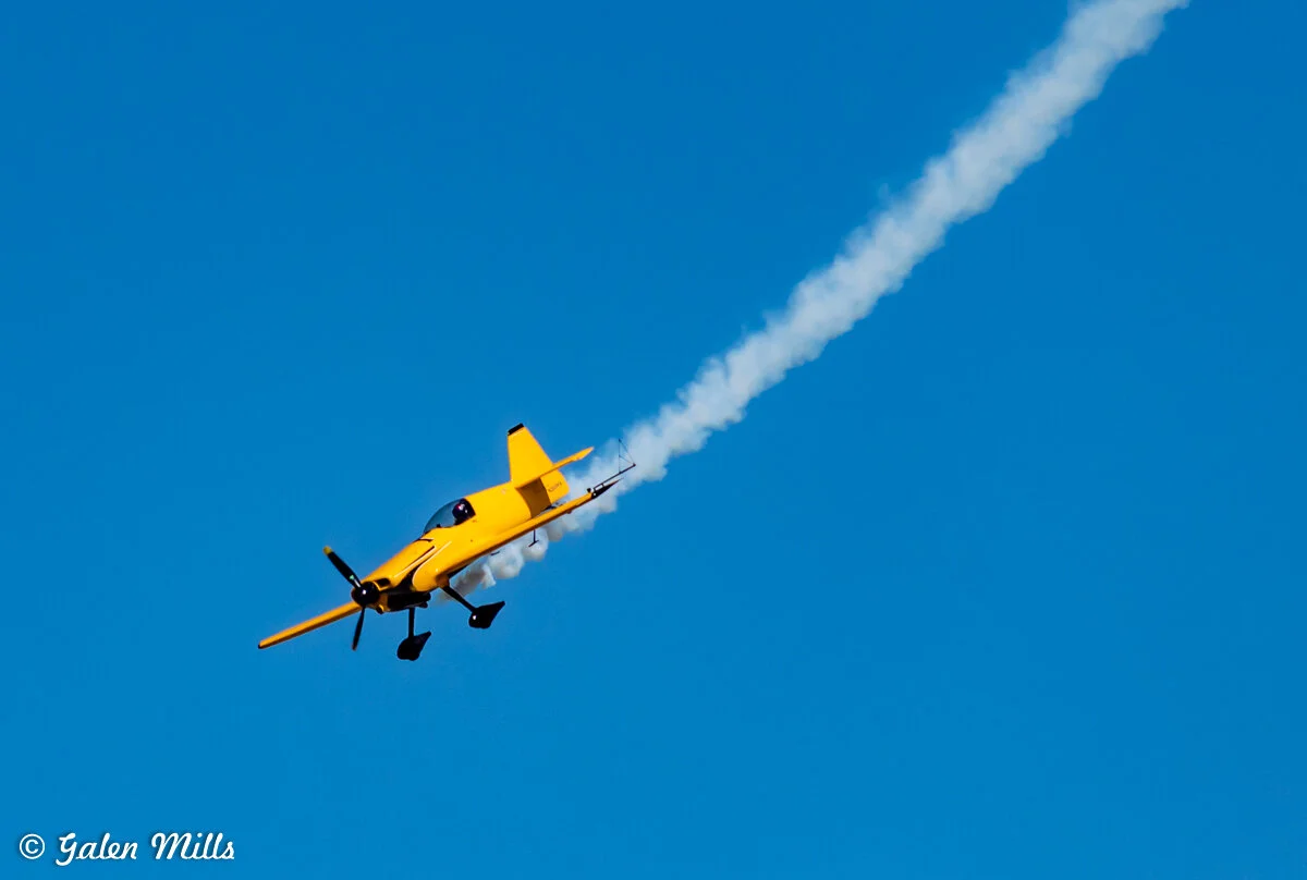 Yellow aerobatic airplane emitting smoke against a clear blue sky.