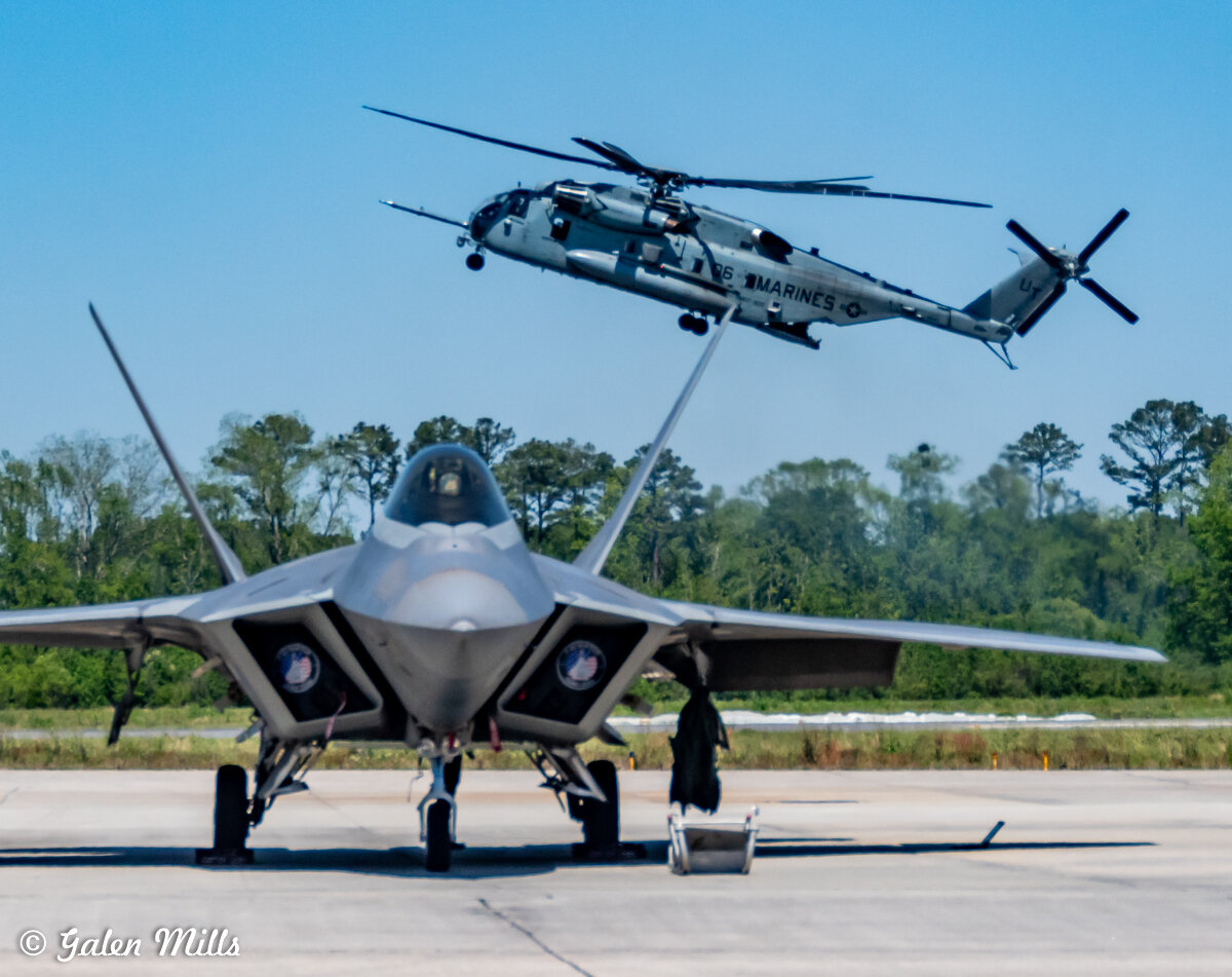 A fighter jet parked on a runway with a helicopter flying in the background.