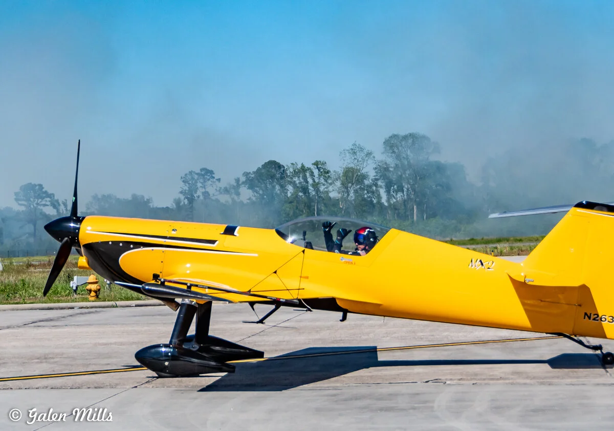 Bright yellow stunt plane on runway with smoke in background and trees in distance.