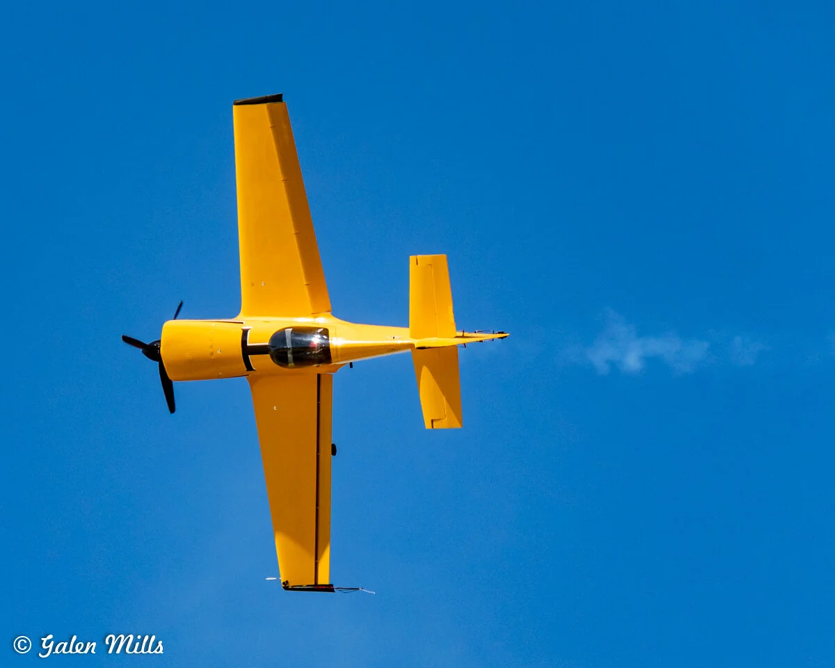 Yellow aerobatic airplane in midair against a blue sky.