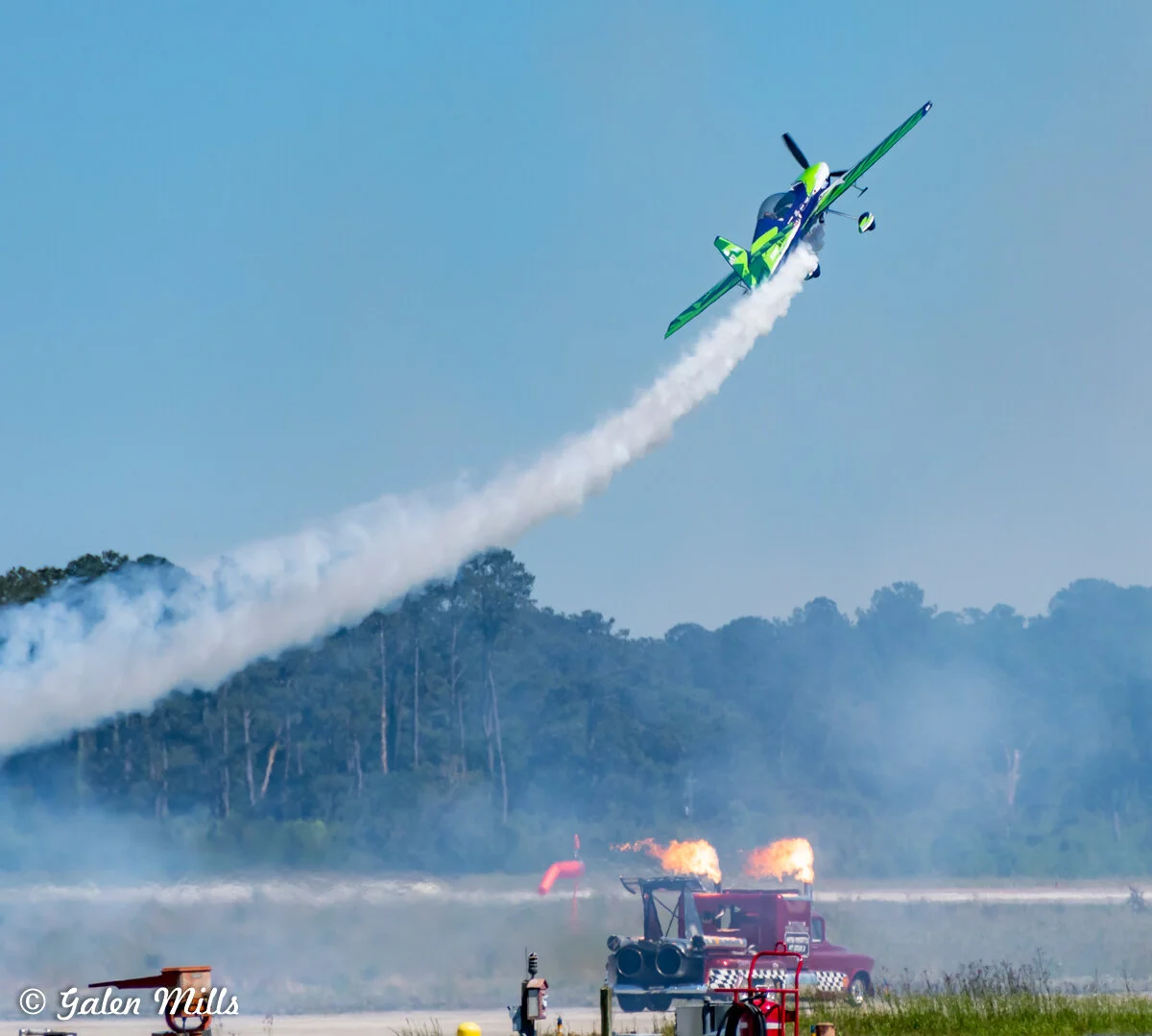 Aerobatic plane performing at airshow with smoke trail and jet-powered truck with flames.