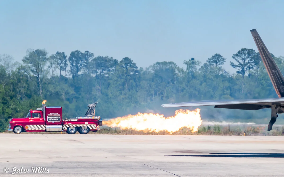 Red truck with flame shooting out the back, parked near an airplane wing on an airfield, with trees in the background.