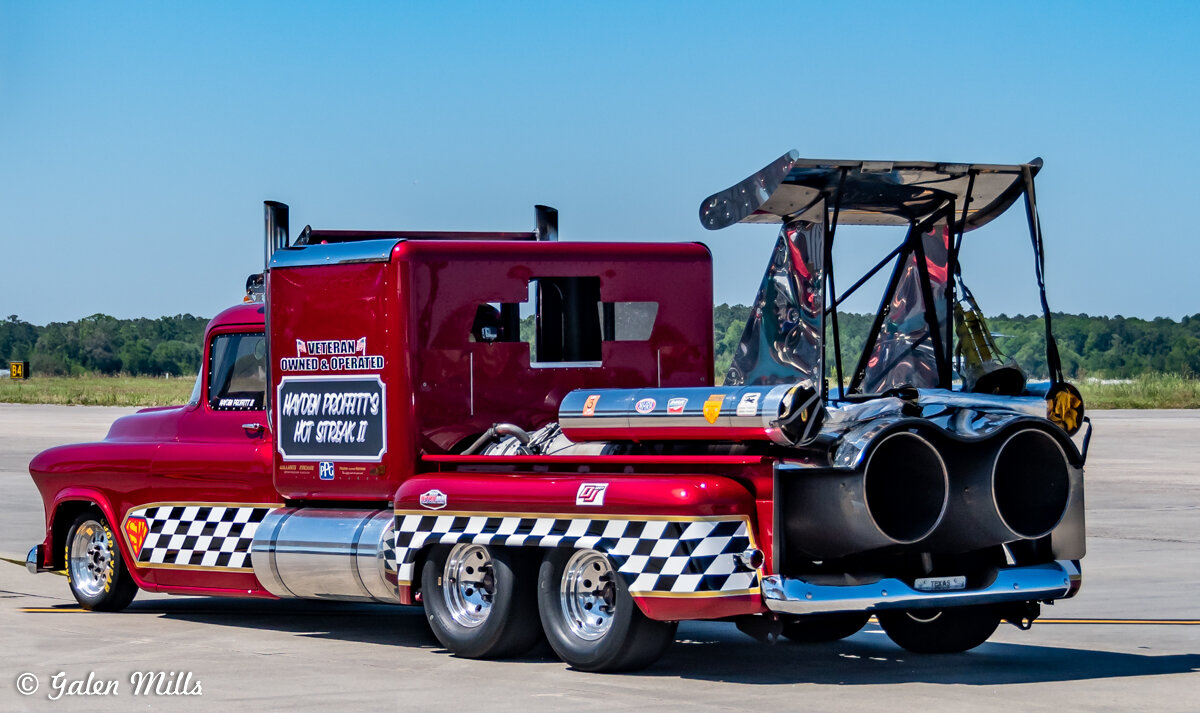 Red custom jet truck with checkered pattern, featuring large rear jet engines, parked on a concrete surface. The truck is labeled as "Veteran Owned & Operated" and "Hot Streak II."