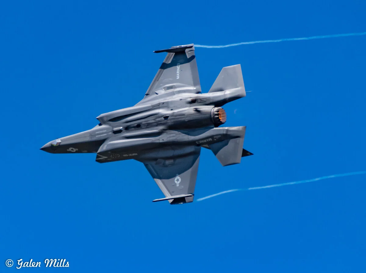 F-35 fighter jet flying in clear blue sky, showing bottom view with visible engine and wings.