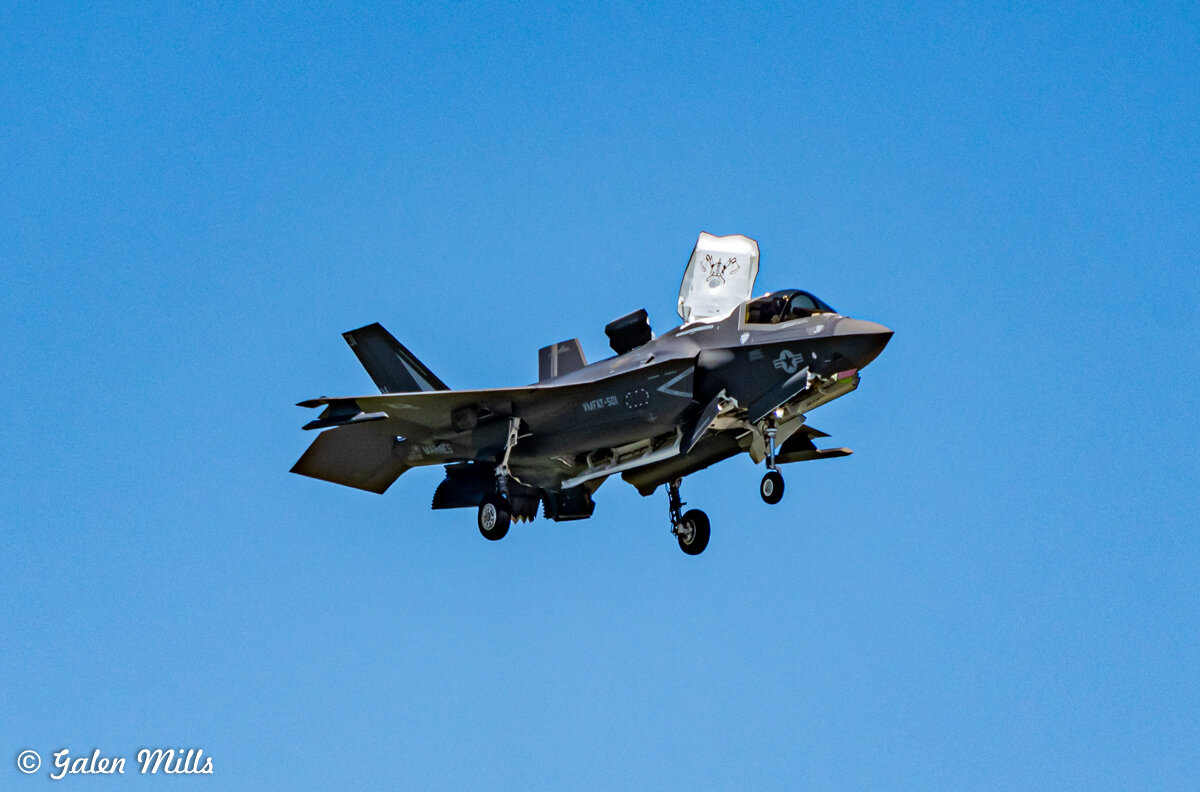 F-35 fighter jet in flight with landing gear extended and vertical lift system engaged against clear blue sky.